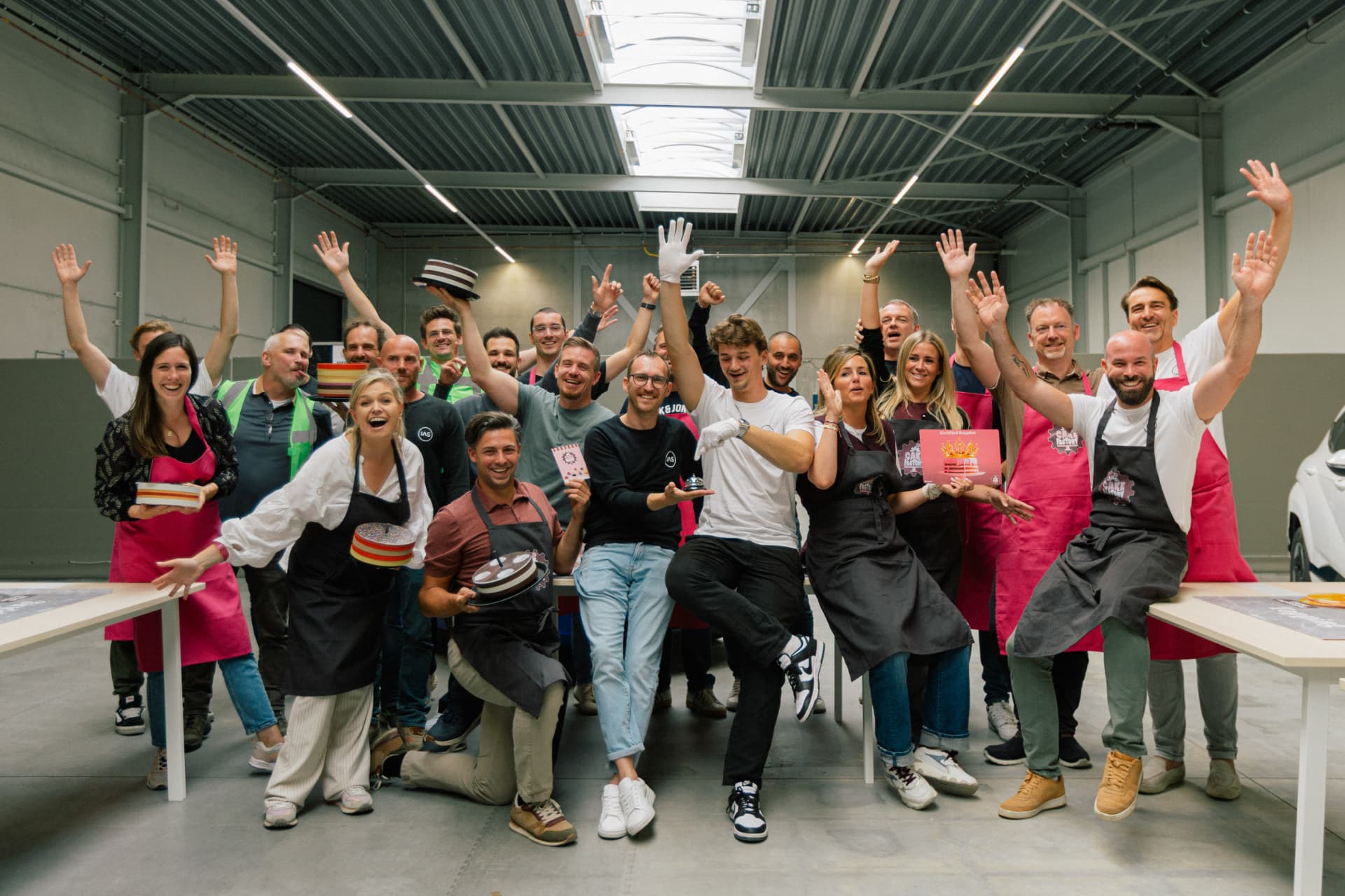 Group portrait of the full IAS team at the Aartselaar HQ, shot against the technical grid wall. Slight smiles, mixed body language, natural light from camera left.